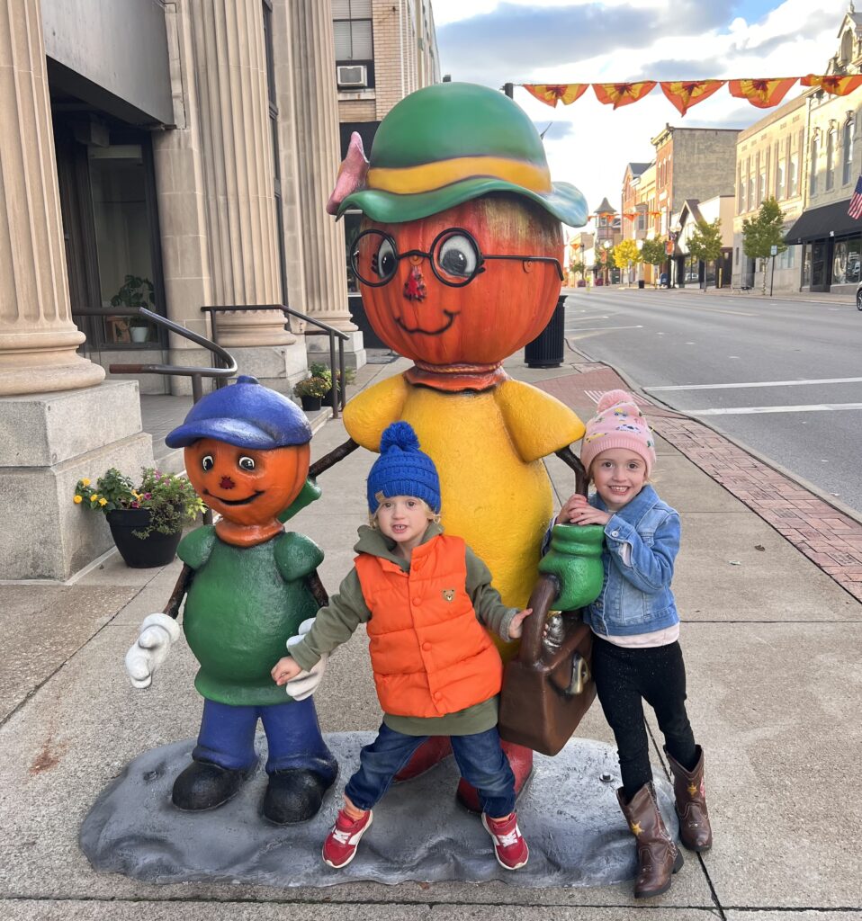 Two children stand beside a pumpkin woman character in bright clothes and a pumpkin child character.
